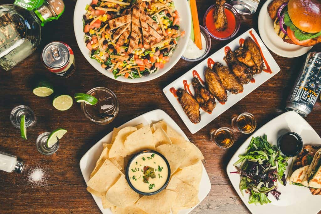 Overhead shot of a lunch spread with salad, wings, chips and dip, tacos, burger, and drinks on a wooden table.